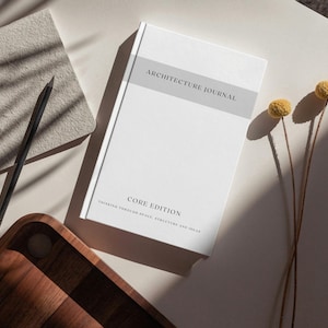 May include: A white Architecture Journal with a gray band and the text "ARCHITECTURE JOURNAL" and "CORE EDITION". A gray notebook, a black pencil, a wooden tray, and two yellow dried flowers are also visible.