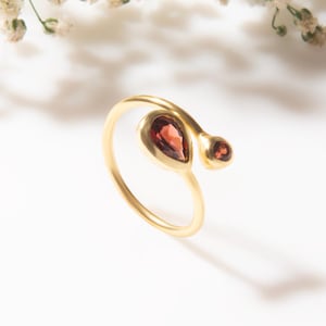 May include: A gold ring with a teardrop-shaped, dark red gemstone and a smaller, round gemstone. The ring is set against a white background with delicate white flowers.
