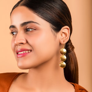 May include: Gold-colored earrings with three circular, textured pendants hanging from a hook. The earrings are worn by a person with dark hair and a brown top, set against a neutral background.