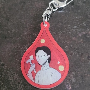May include: Red teardrop-shaped keychain with a silver clasp. The keychain features a pixelated illustration of a person holding a hammer. The background is red with yellow dots.
