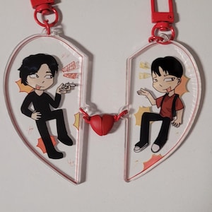 May include: Two-piece heart-shaped keychains with cartoon illustrations of two people. Each keychain half has a red clasp. A small red heart connects the two halves. The characters are in black and red outfits.