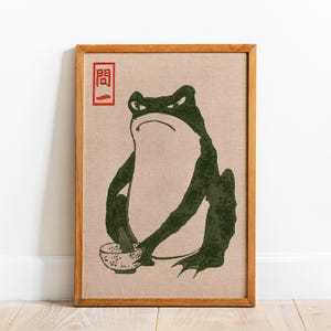 May include: Framed art print featuring a green frog holding a bowl, with a red Japanese character in the upper left corner. The frog has an angry expression. The artwork is set against a beige background and framed in wood.