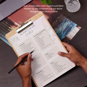 Peut inclure: Un presse-papiers en bois avec une liste de contr&ocirc;le en allemand intitul&eacute;e "KLINIKTASCHE". La liste comprend des sections pour les documents, le s&eacute;jour de la m&egrave;re, la naissance, l'allaitement, les extras et les articles pour b&eacute;b&eacute;. Un stylo est tenu au-dessus de la liste.