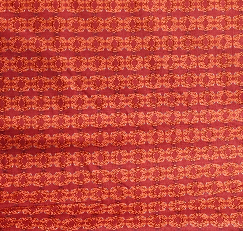 Op de afbeelding: Rode stof met een herhalend patroon van oranje en witte geometrische vormen.