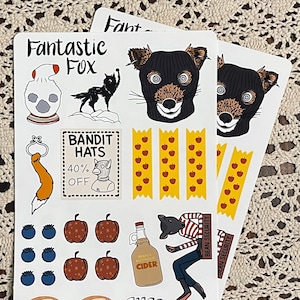 May include: A set of stickers featuring a fox wearing a black ski mask, apples, a fox tail, a glass jar, a fox wearing a striped shirt, a bottle of cider, pumpkins, blueberries, and a sign that says "Bandit Hats 40% Off".