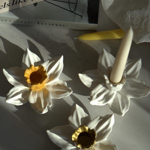 May include: Three ceramic candle holders shaped like white daffodils with yellow and gold centers. One holder contains a tapered cream candle. The base of the holders features sculpted petals, and the center forms a cup for a candle.