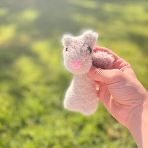 May include: A small, handmade, light gray bunny plushie with a pink nose and black eyes. The bunny has a fluffy texture and is held in a hand against a blurred green background. The plushie is a cute, handcrafted item.