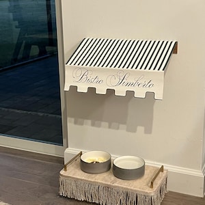 May include: A pet feeding station with a black and white striped awning that reads "Bistro Simberto." Below, two gray bowls sit on a fringed, rectangular tray with gold handles. The bowls are for food and water.