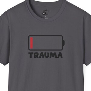 May include: Dark gray t-shirt featuring a red and black graphic of a low battery with the word "TRAUMA" below. The shirt has short sleeves and a crew neck.