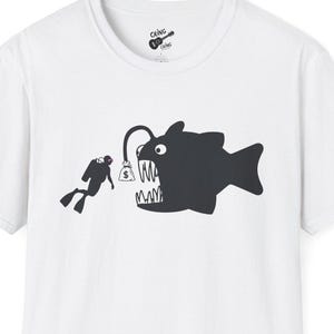 Puede incluir: Camiseta blanca con una ilustración negra de un buzo y un rape. El rape tiene la palabra "MA" en la boca. El buzo nada hacia el pez. La camiseta tiene cuello redondo y mangas cortas.