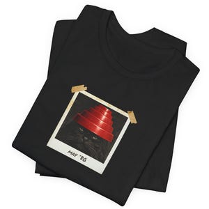 Op de afbeelding: Zwart T-shirt met een foto in Polaroid-stijl van een zwarte kat met een rode, gelaagde hoed. De foto is met bruine tape op het shirt geplakt en heeft de tekst "MAY '80". Het shirt is opgevouwen.