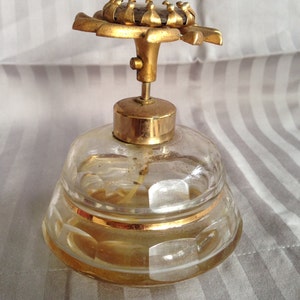 May include: A vintage glass perfume bottle with a gold-toned flower-shaped stopper. The bottle has a clear glass body with a textured design and a gold band around the top.