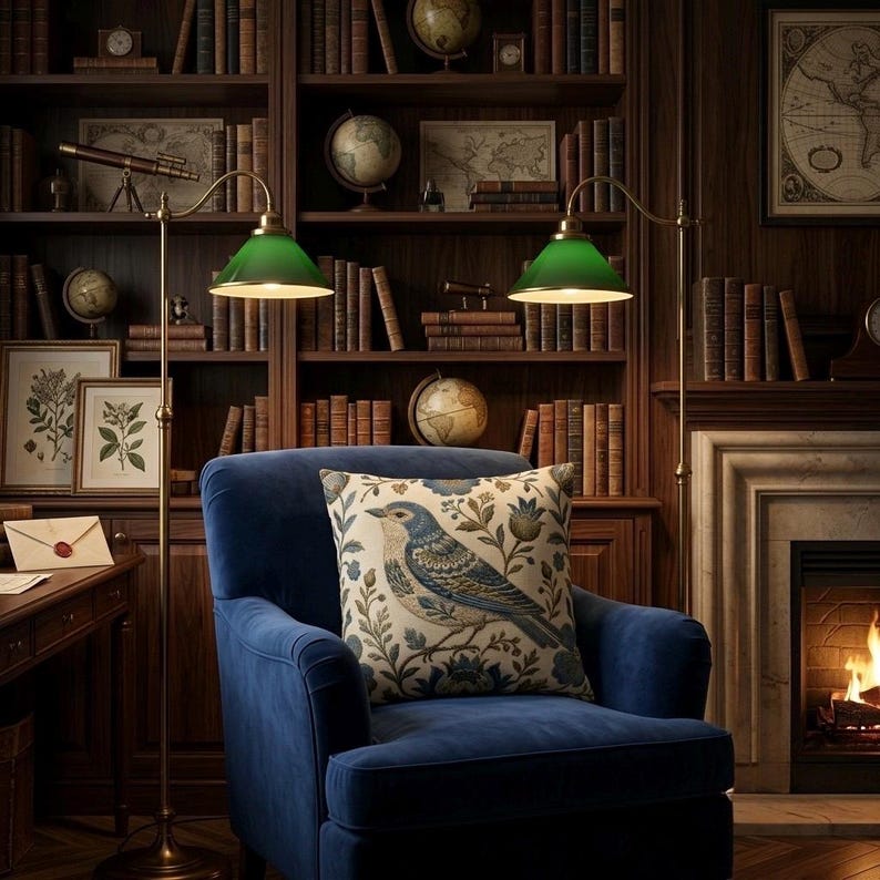 May include: A deep blue velvet armchair featuring a square throw pillow with a detailed bird and floral pattern. Two brass floor lamps with green glass shades stand on either side of the chair, illuminating the seat in a library setting.