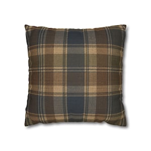 May include: A square throw pillow featuring a plaid pattern in shades of dark brown, tan, and muted blue. The fabric has a visible woven texture with intersecting horizontal and vertical lines forming a classic grid design across the entire surface.