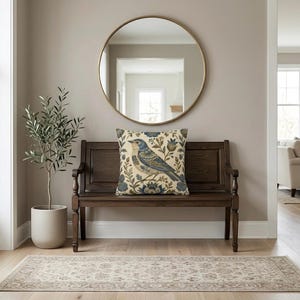 May include: A square throw pillow featuring a detailed illustration of a blue bird perched among stylized floral and botanical elements in shades of blue, brown, and cream, set against a light cream background with a textured fabric appearance.