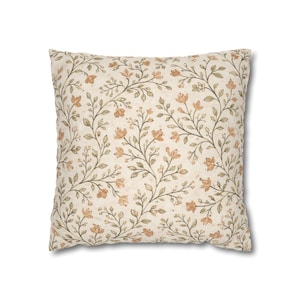 May include: Square throw pillow with a cream-colored background and a repeating floral pattern. The design features delicate peach-colored flowers and green leaves on winding stems, creating a vintage aesthetic. The pillow is a decorative home accent.