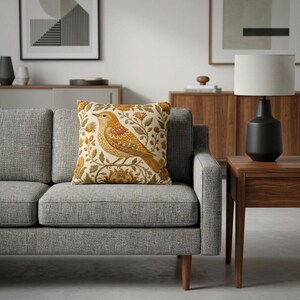 May include: A square throw pillow featuring a cream-colored background with a detailed illustration of a golden-yellow bird perched among stylized orange and green floral vines and leaves, centered on a gray upholstered sofa cushion.