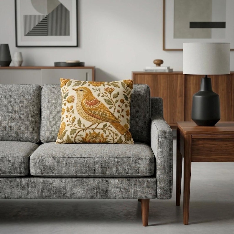 May include: A square throw pillow featuring a cream-colored background with a detailed illustration of a golden-yellow bird perched among stylized orange and green floral vines and leaves, centered on a gray upholstered sofa cushion.