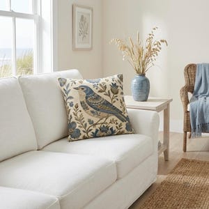 May include: A square throw pillow featuring a woven pattern of a blue and tan bird perched among blue flowers and green foliage. The pillow is placed on a white sofa next to a blue ceramic vase filled with dried wheat stalks on a side table.
