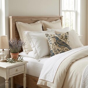 May include: A wooden headboard with a light beige duvet and matching pillow shams. A square decorative pillow features a blue bird and floral pattern. A small terracotta pot with lavender sits on a white wooden nightstand next to a table lamp.