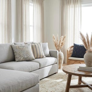 May include: A light gray sectional sofa with a floral patterned pillow and a striped pillow, a round wooden coffee table, a rattan chair with a dark blue pillow, and two large ceramic vases filled with dried pampas grass in a bright room.