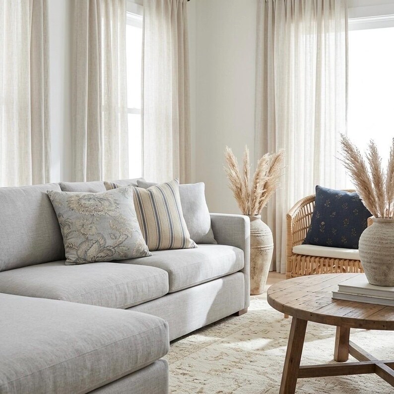 May include: A light gray sectional sofa with a floral patterned pillow and a striped pillow, a round wooden coffee table, a rattan chair with a dark blue pillow, and two large ceramic vases filled with dried pampas grass in a bright room.