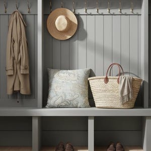 May include: A beige trench coat hangs on a hook next to a straw hat. A square pillow with a light blue and beige floral pattern sits on a bench beside a woven straw basket with brown leather handles and a small gray fringed scarf draped over it.