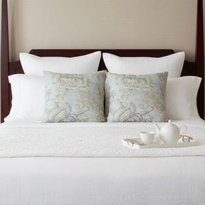 May include: A bed featuring a dark wood headboard, white quilted bedding, and two square throw pillows with a light blue and beige floral pattern. A white ceramic teapot and a matching teacup sit on a white rectangular tray on the bed.