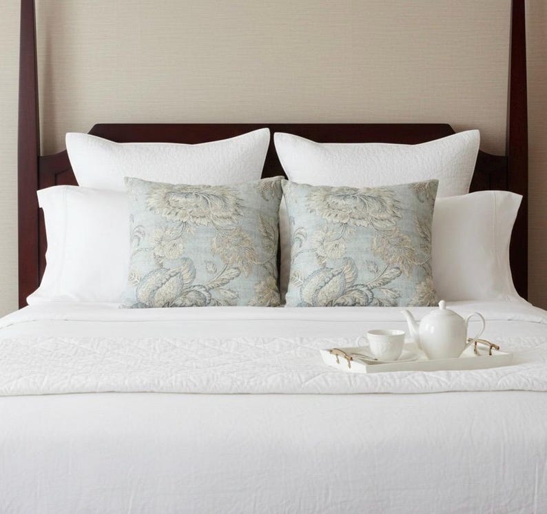 May include: A bed featuring a dark wood headboard, white quilted bedding, and two square throw pillows with a light blue and beige floral pattern. A white ceramic teapot and a matching teacup sit on a white rectangular tray on the bed.