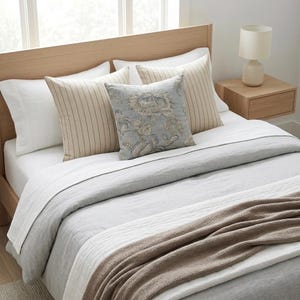 May include: A bed featuring a light wood headboard, white textured pillows, two beige striped square pillows, and one square pillow with a blue and beige floral pattern. The bedding includes a light gray duvet and a folded brown knit throw blanket.