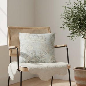 May include: A square throw pillow featuring a light blue and beige floral pattern. The pillow is placed on a wooden chair with a woven cane backrest and black metal armrests, with a white fabric draped over the seat of the chair.