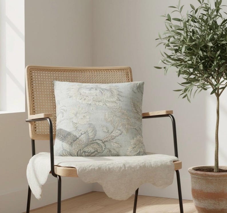 May include: A square throw pillow featuring a light blue and beige floral pattern. The pillow is placed on a wooden chair with a woven cane backrest and black metal armrests, with a white fabric draped over the seat of the chair.