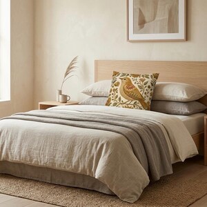 May include: A square throw pillow featuring a detailed illustration of a brown bird perched on a branch, surrounded by floral and botanical motifs in shades of gold, green, and rust on a cream-colored background, placed on a bed with neutral bedding.