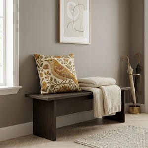 May include: A dark brown wooden bench featuring a square throw pillow with a bird and floral pattern in shades of gold, orange, and green. A folded cream-colored knit throw blanket rests on the bench next to the pillow, with two umbrellas nearby.
