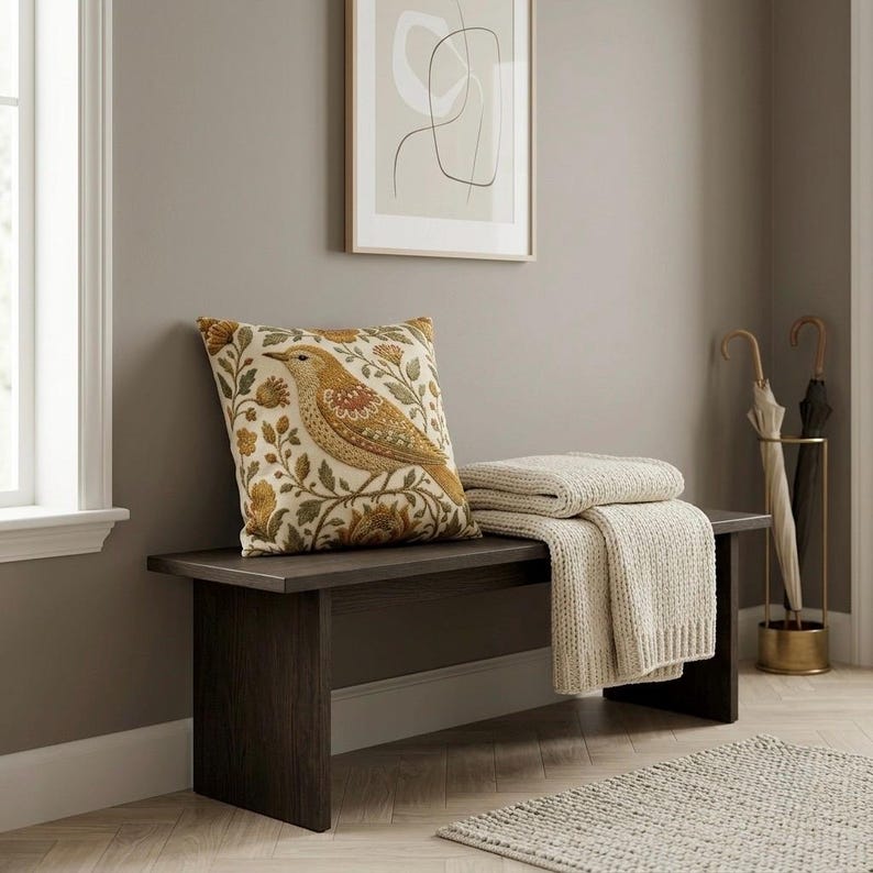 May include: A dark brown wooden bench featuring a square throw pillow with a bird and floral pattern in shades of gold, orange, and green. A folded cream-colored knit throw blanket rests on the bench next to the pillow, with two umbrellas nearby.