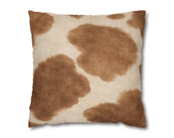 May include: Square decorative pillow with a cowhide pattern. The pillow features large, irregular brown spots on a cream-colored background. The pillow is square and appears to be made of a soft fabric.