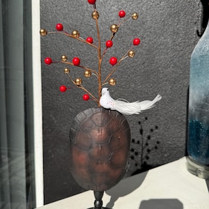May include: A decorative centerpiece featuring a dark brown, turtle-shell-shaped base with a black stand. A branch with red and gold spherical ornaments and a white dove figurine adorns the top. The piece is set against a textured gray background.