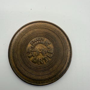 May include: A bronze-coloured, circular object with concentric grooves, resembling a vinyl record. The centre features the text "STAPES RECORDS TURTLE" in a stylized font, with a turtle graphic. The object has a worn, vintage appearance.