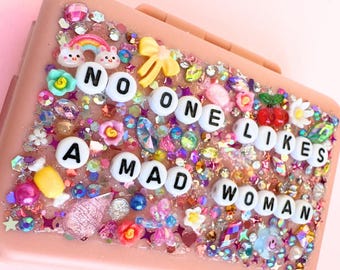 Puede incluir: Una caja rectangular rosa decorada con cuentas de colores, pedrería y pequeños abalorios. La caja tiene la frase "NO ONE LIKES A MAD WOMAN" escrita con cuentas de letras blancas. La caja está adornada con flores, arcoíris y otros elementos decorativos.