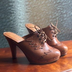 Könnte beinhalten: Ein Paar braune Lederclogs mit hohen Absätzen. Die Clogs haben ein Schnürdesign und dekorative Nähte. Die Absätze sind aus Holz und gebogen. Die Schuhe werden auf einer Holzoberfläche präsentiert.