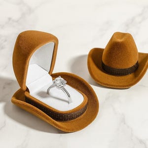 May include: A brown cowboy hat-shaped ring box with a silver ring featuring a large, clear gemstone. The ring box is open, revealing a white interior. A second, closed hat-shaped box is also present.