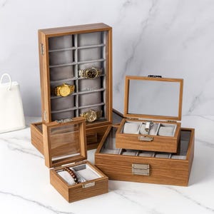 Op de afbeelding: Houten horloge- en sieradendozen in verschillende maten. De dozen zijn gemaakt van lichtbruin hout met grijze fluwelen interieurs. Sommige hebben glazen deksels en bevatten horloges en een armband.