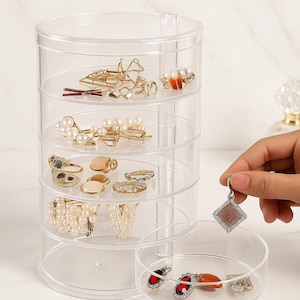 Pode incluir: Um organizador de joias transparente de vários níveis preenchido com vários acessórios. O organizador contém anéis, brincos, grampos de cabelo e outras joias. Uma mão segura um pingente quadrado com uma moldura prateada e um centro marrom.