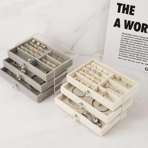 May include: Two clear acrylic jewelry organizers with multiple drawers. One is gray, the other is cream-colored. Each drawer is lined with velvet and contains rings, earrings, bracelets, and necklaces. The organizers have clear, decorative knobs.