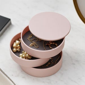Puede incluir: Un organizador de joyas rosa claro de tres niveles con tapa a juego. Los compartimentos abiertos revelan collares, anillos y pendientes de perlas doradas. El interior de cada compartimento es de color gris oscuro.