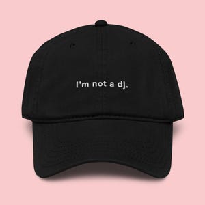 Puede incluir: Gorra de béisbol negra con visera curva y correa ajustable. La gorra presenta el texto bordado "I'm not a dj." en blanco en el panel frontal. Hecha de tela suave y duradera, es un accesorio casual.
