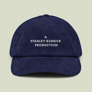 May include: A navy blue corduroy baseball cap featuring the embroidered text "A STANLEY KUBRICK PRODUCTION" in white. The cap has a curved peak and an adjustable strap. A classic, stylish accessory for everyday wear.