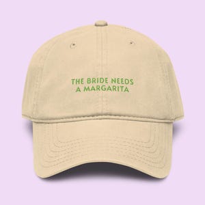 Może przedstawiać: Beżowa czapka bejsbolowa z zielonym napisem "THE BRIDE NEEDS A MARGARITA". Czapka ma zakrzywiony daszek i regulowany pasek z tyłu. Kapelusz jest wykonany z miękkiego, trwałego materiału.