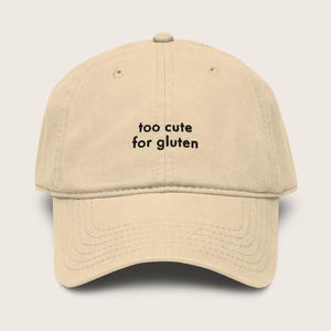 Peut inclure: Casquette de baseball beige avec l'inscription "too cute for gluten" en noir. La casquette a une visière incurvée et un bouton sur le dessus. Le chapeau est fait d'un tissu doux et texturé.