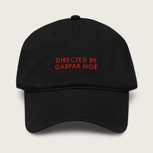 May include: A black baseball cap featuring the text DIRECTED BY GASPAR NOÉ embroidered in red thread on the front panel. The cap has a structured crown, a curved brim, and a button at the top center, showcasing a clean and simple design.